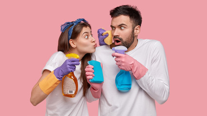 Shot of woman and man housekeepers hold sprays and sponges, lovely female folds lips for kissing...