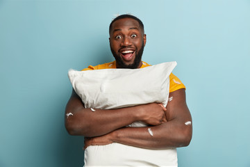 Isolated shot of dark skinned young man with optimistic gaze carries pillow, has fun in bedroom,...