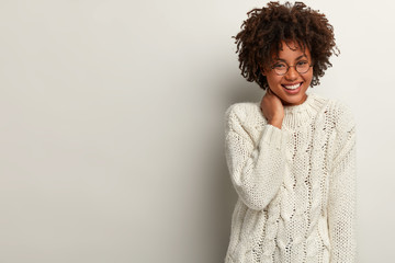 Glad laughing curly woman giggles as heard funny joke, wears white sweater, keeps hand on neck, expresses positive emotions, stands indoor, blank space right for your advertisement. Happiness - obrazy, fototapety, plakaty
