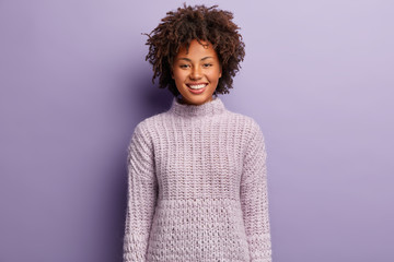Portrait of happy Afro American woman with curly hair, smiles gladfully, has healthy skin, wears knitted purple sweater, poses indoor, laughs at something funny. Monochrome. People and positiveness