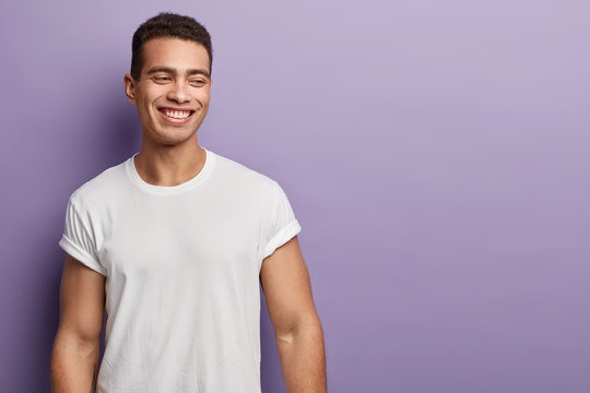 Indoor shot of handsome muscular guy with positive expression, wears casual mock up white t shirt for your promotion, stands against purple background, free space aside. People and emotions.