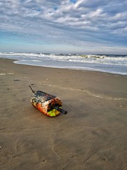 Obraz premium Float from a crab trap washed ashore on a sandy beach. Waves crash onto the shore in the background and cloudy sky. Fishing industry trash and debris. Coastal pollution.