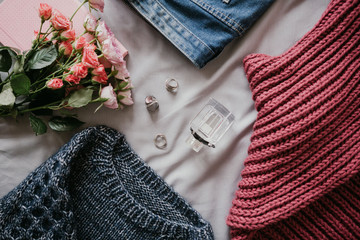 Clothing layout: perfume, jeans, sweater, scarf, rings and flowers on a gray fabric background