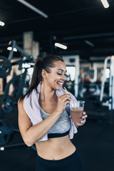 Young attractive woman after successful workout in modern fitness gym holding glass of protein shake and drinks with drinking straw.