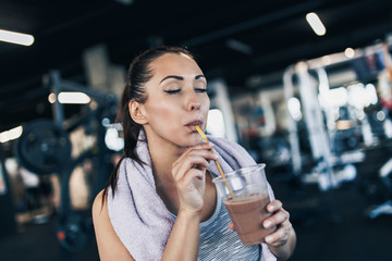 Young attractive woman after successful workout in modern fitness gym holding glass of protein shake and drinks with drinking straw. - obrazy, fototapety, plakaty