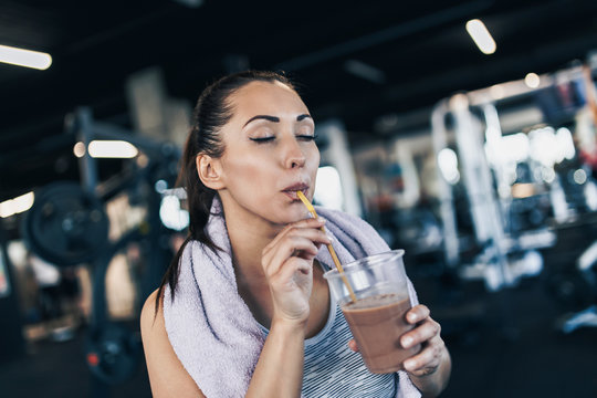 Young Attractive Woman After Successful Workout In Modern Fitness Gym Holding Glass Of Protein Shake And Drinks With Drinking Straw.