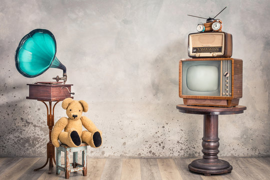 Retro TV receiver and old broadcast radio from circa 50s, alarm clock on wooden table, outdated gramophone, Teddy Bear toy sitting on stool front concrete wall background. Vintage style filtered photo