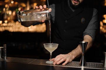 Male bartender pouring a delicious cocktail from the measuring glass cup through the strainer