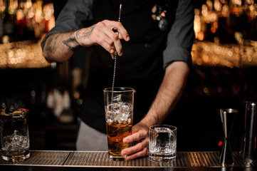 Bartender stirring many ice cubes in the tall glass with a brown cocktail with a spoon