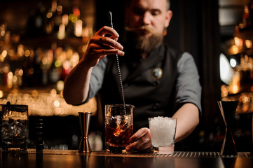 Bartender with a beard stirring alcohol with an ice cubes in the tall measuring glass cup with a...