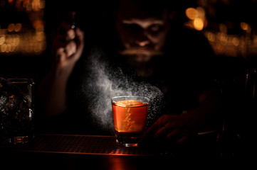 Bartender spraying to the cocktail glass with one big ice cube in the dark