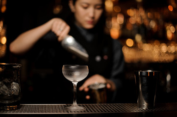 Cold cocktail glass on the blurred foreground of bartender girl placing an ice cubes to the cup