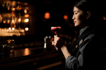 Female bartender holding a smooth crimson cocktail in the glass with a one pink rose bud as a decor