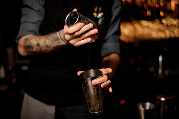 Bartender with a beard and tattoo on hands making cocktail in the steel shaker