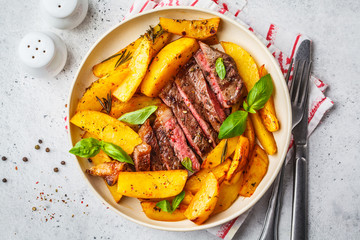 Grilled beef steak with potatoes and basil in a white plate, top view.