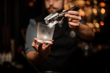 Professional bartender holding in hand an ice cube in tweezers putting it on the a cold matte cocktail glass
