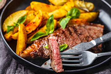 Grilled beef steak with potatoes and basil in a black plate on dark background.