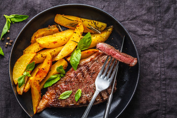 Grilled beef steak with potatoes and basil in a black plate on dark background, top view.