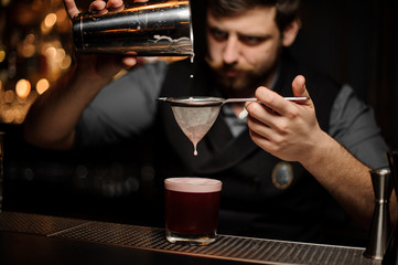Bartender with a beard pouring a smooth pink cocktail through the sieve to the glass from the shaker