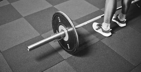 Fototapeta na wymiar Woman trains in the gym with a barbell. Powerlifting. Heavy barbell on the floor and female legs. Black and white sport photo.