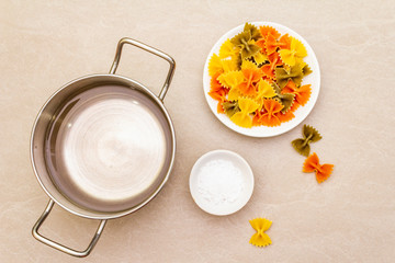 Raw farfalle with water in pot and salt. Multicolored pasta in white plate on stone background. Preparing for children, top view, close up