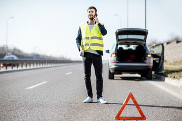 Man in vest calling road assistance standing near the broken car on the highway