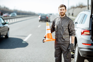 Portrait of a happy road assistance worker in uniform standing with cone near the car on the roadside