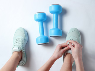 Legs of young sports woman, plastic dumbbells. Woman tying shoelace of sports shoes on a white background. Top view. Minimalism