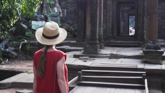 Attractive young woman walking through one of the most famous temples in Angkor, the Ta Prohm. Shot from the back of a woman in the red dress walking into the Ta Prohm, Angkor, Cambodia. 