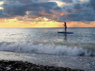 Young female paddling on SUP board in the sea at amazing dark sunset and waves. Sup surfing woman silhouette - active recreation in nature. 