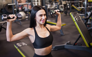 Fototapeta premium Cheerful athlete woman smilling and doing dumbbell press up exercise on sitting on bench in gym
