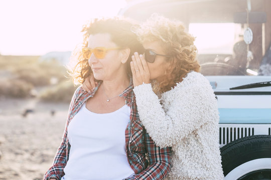 People Traveling Together - Couple Of Caucasian Curly Friends Women Whispering At The Ear Some Secrets With Sunny Sunset In Backgorund - Old Vintage Van For Alternative Travel Vacation Concept