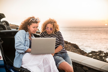 Couple of people women friends use together a laptop computer sitting on the back of convertible car and sunset on the ocean in background - work everywhere and travel vacation plan concept