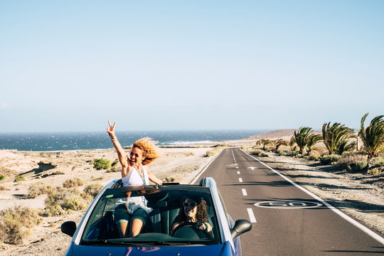 Happy cheerful couple of young caucasian crazy woman with curly hair enjoy the travel and vacation staying outside the roof of a convertible car with long road and ocean in background - joyful