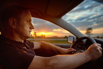 Fototapeta premium man sitting behind the wheel of car during his anticipated travel on summer vacations, man riding in automobile at the countryside with beautiful evening sky and sun rays. Lifestyle and travel concept