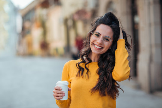 Beautiful casual brunette with coffee to go in the city street, smiling at camera.