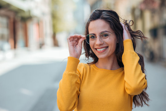 Headshot of a positive young woman with eyeglasses in the city, smiling at camera.