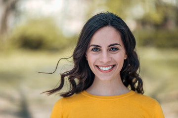 Headshot of a young brunette woman with a toothy smile on a light outdoor background.