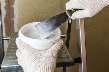 Male gloved hand holds a construction spatula over a putty jar against a wall with a smeared crack....