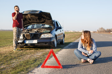 Outdoor shot of couple near his brocken car, red triangle as warning sign, male stands in front of opened hood and calls tow truck, female sits on road with crossed legs, waits solving problem.