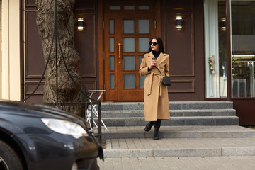 Fototapeta na wymiar Full length photo of beautiful brunette female standing near city building, drinking hot takeaway coffee, going to her car, looks aside, wearing beige coat, boots, sunglasses, spends free time.
