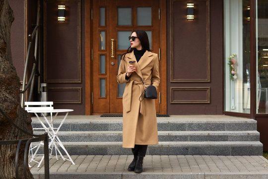 Beautiful young brunettee woman in modern beige coat, holding disposable takeaway cup and standing against urban city building, looks thoughtfuly aside, has dating, waiting for her boyfriend.