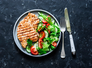 Grilled turkey chops and tomatoes, cucumbers, greens salad on a dark background, top view. Healthy...