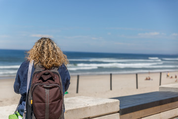 A tourist woman with a backpack on her back, placidly observes a calm sea on a beach, on a sunny day