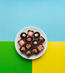 Beautiful chocolates of different shapes and fillings lie in a white plate against the background of colored paper. Copy space, vertical. Yellow, green, blue color.