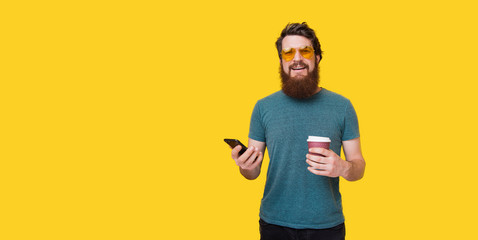 Handsome bearded man holding coffee and phone in hands.  Hipster in stylish yellow glasses over isolated background