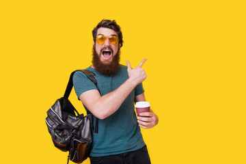 Excited bearded man with stylish eyeglasses, pointing away while holding a cup with coffee on...