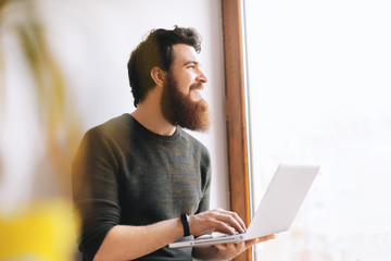 Handsome cheerful bearded man sitting near window, thinking and looking forward while using laptop