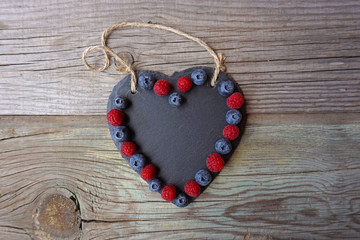 Raspberries and blueberries atop the black stone heart decoration on an old wooden surface. Fresh berries. Romantic concept. Summer food. 