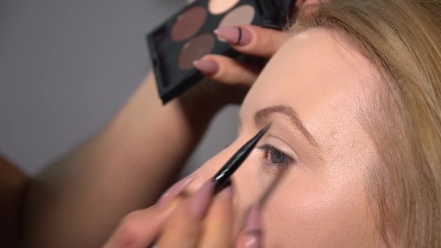 Beauty saloon. Young beautiful girl model is sitting in the chair. Makeup artist makes girl makeup. Makeup artist applies eyebrow shades on model's face.